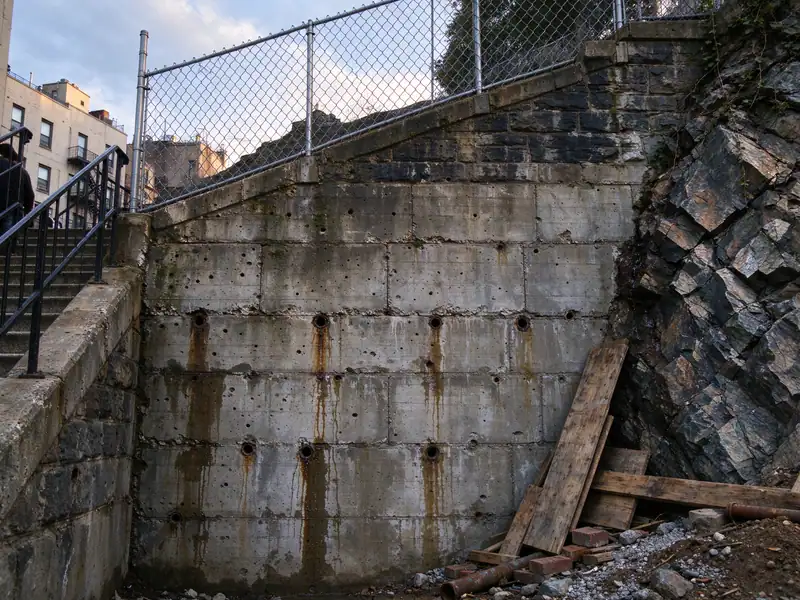 Very tall weathered concrete retaining wall with water staining beside a public stairway in upper Manhattan
